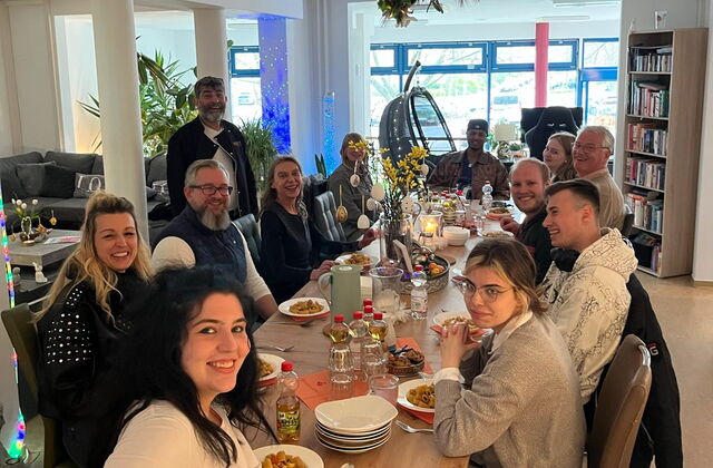 Langer gedeckte Tisch mit 12 Personen. Gemeinsames Abendessen. Alle lächeln in die Kamera.