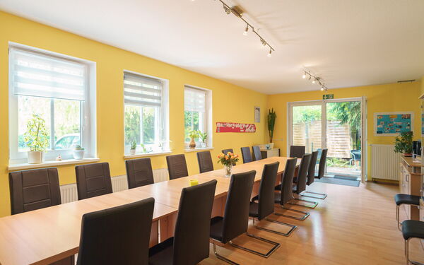 Großer Speiseraum. Langer Tisch mit 17 Stühlen. Die Wandfarbe ist Gelb, Fenster und Terrassentür lassen viel Licht in den Raum.