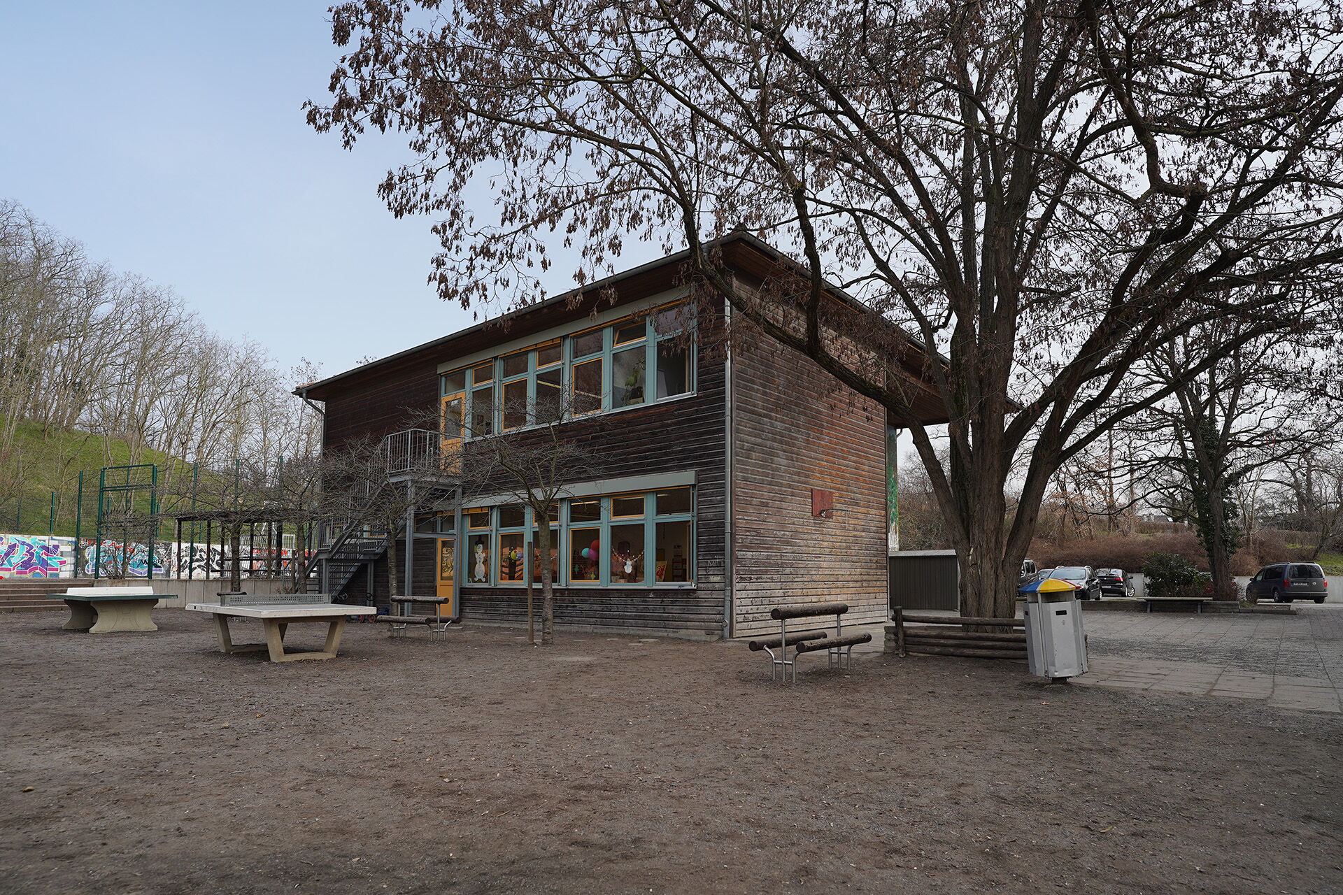 Außenbereich mit Kitagebäude. Draußen, großer Baum neben dem Gebäude und zwei Tischtennisplatten davor.