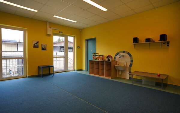 Großer Raum, gelbe Wände, Teppich blau. An der Wand ein Regalbrett wo eine Musikanlage mit zwei Boxen steht.