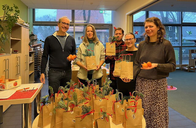 Im Raum. Runder Tisch mit vielen Nikolaus-Papier-Tüten, jede mit Weihnachtsaufkleber und Tannenzweigen verziert. Dahinter stehen zwei Männer und drei Frauen die lächeln und Tüten oder Mandarinen in den Händen halten.