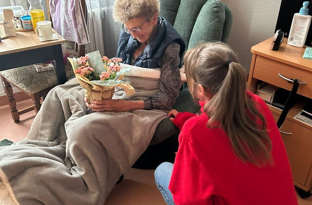 Bewohnerzimmer. Eine ältere Frau mit eingewickelten Blumentopf in den Händen sitzt in einem grünen Sessel mit eine grauen Decke auf den Beinen.