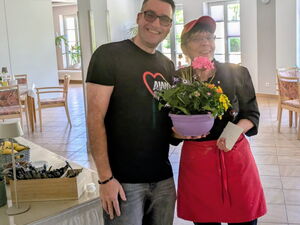 Cafeteria: Mann und Frau stehen im Zentrum und lächeln in die Kamera. Die Frau hält einee lila Blumenschale in der Hand.