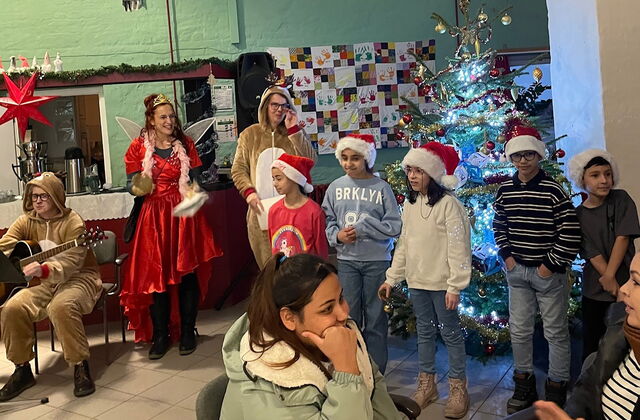 Weihnachtlich geschmückter Raum. 5 Kinder mit stehen vor einem festlichem Weihnachtsbaum. Eine Person im Renntierkostüm sitzt mit einer Gitarre in den Händen auf einem Stuhl. Ein Frau im roten Feenkostüm strahlt und klatscht. Alle sind sehr fröhlich.