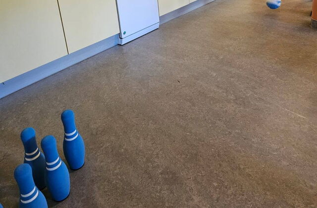 Küchenzeile.   Daneben stehen blaue Kegel. Ein ältere Mann am rechten Bildrand kullert ein Ball in Richtung Kegel.
