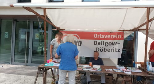 Überdachter Info-Stand des Ortsvereins. Zeitungen, Flyer, Broschüren liegen auf dem Tisch. Zwei Menschen im Stand informieren eine Person die davor steht.