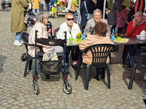 Sonne scheint, Senioren sitzen an einen geschmückten Tisch. Dahinter Markttreiben.