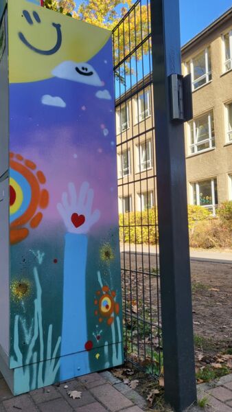 Eine Art Container von der Seite bunt bemalt mit lächelnder Sonne, Wolke, bunten Blumen. Der Container steht an einem hohen Zaun.