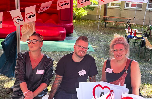Drei Personen sitzen auf einer Bank im AWO-Infostand. Zwei Frauen, ein Mann. Sie lächeln fröhlich in die Kamera.