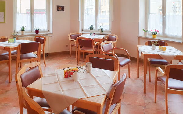Ein Speisesaal. 4 kleine vierer Tische im Raum verteilt. Helle Raum, Fenster lassen viel Licht herein.