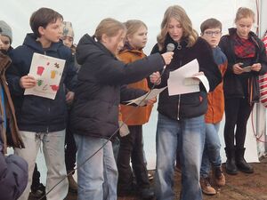 Circa. 12 Kinder die etwas vortragen. Ein Mädchenn hält einem anderen Mädchen ein Mikrofon hin. Einige haben Zettel in den Händen.
