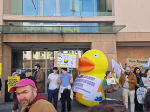 Viele Menschen vor den Türen eines Gebäudes. Eine große gelbe Ente mit einer Aufschrift. Ringherum Menschen mit Schildern und AWO Fahnen. Auf einem Schild steht: DEMO gegen Kürzungen.