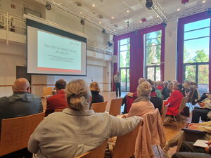 Großer Saal. Viele Stuhlreihen wo Menschen sitzen, Leinwand forne, ein Redner mit Mikrofon steht vorn.