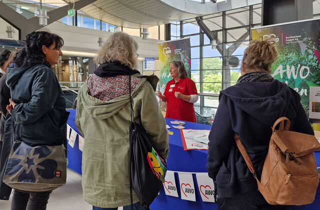 In den Bahnhofspassagen. Stand der AWO mit mehren Personen die sich informieren davor. Frau im roten AWO-T-Shirt steht hinter dem Tisch mit Info-Material.
