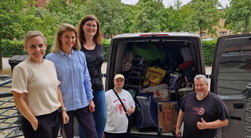 3 Frauen stehen im Vordergrund auf einer Erhöhung. Dahinter und unten auf Straße ein vollgepackter Transporter von hinten, beide Türen geöffnet. Zwei Männer stehen vor dem Laderaum machen ein Daumen-Hoch.