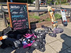Draußen. Ein Schild: Skatebord und Urban Art Workshops.  3 Skateboards lehnen an einer Stand und Schutzkleidung wie Knieschützer liegen wild verteilt auf dem Boden,