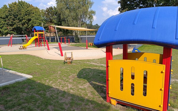 Spielplatz, Sandkasten, Schaukel, Klettergerüst mit Rutsche. Wiese Bäume, die Sonne scheint.