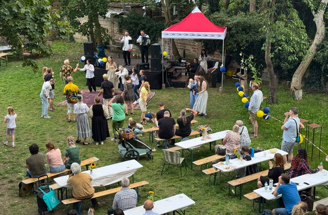 Von schräg-oben: Eine Wiese, mit Bänken und Tischen, eine kleine Bühne und roter Pavillon. Eine Band spielt auf der Bühne, Gäste tanzen davor.