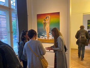 Ausstellungsraum. Drei Frauen stehen vor einem Bild an der Wand. Das Bild zeigt eine nackte Frau mit langen Haaren am Strand stehend. Die Hintergrund ist Regenbogen-farbig.