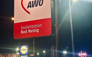 beleuchtetes Schild mit dem AWO Logo, im Hintergrund Straßenlaternen, Hauptstraße, beleuchtetes Schild von Lidl und Parkplatz von Rewe, Himmel ist dunkel