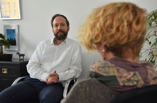 Ein Büroraum. Mann sitzt einer Frau gegenüber. Die Kamera ist auf den Mann gerichtet, die Frau nur von hinten im Bild. Mann trägt weißes langärmliches Hemd, hat einen Vollbart und dunkle Haare.
