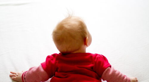 Ein Baby im roten Strampelanzug und hellen Haaren, welches auf dem Boden krabbelt. Von hinten oben.