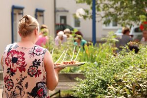 Eine Frau trägt einen einem Teller mit Kuchenstücken. Die Sonne scheint, es ist warm. Die Frau trägt ein ärmelloses Shirt mit großen Blüten und hat die langen blonden Haare hochgesteckt.