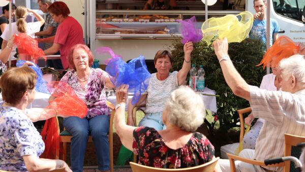 Seniorinnen sitzen in einem Stuhlkreis und schwenken farbige Tücher in der Luft. Die Tücher sind blau, lila, gelb und rot.