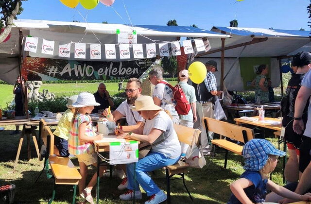 Vor Marktständen sind Tische und Bänke aufgestellt, an denen 2 Kinder und 2 Erwachsene sitzen und die Kinder malen Bilder. Der Stand ist mit Luftballons und Wimpeln geschmückt. Die Sonne scheint.