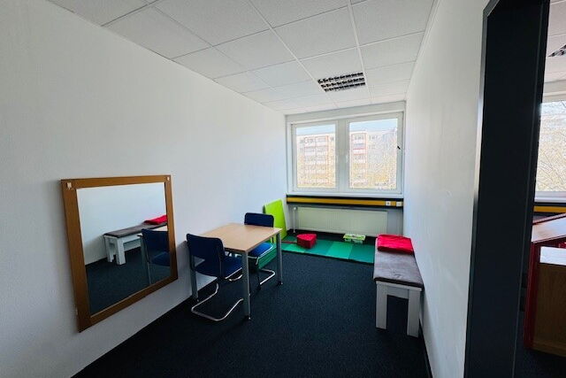 Ein sparsam ausgestatteter Raum mit einem Tisch und zwei Stühlen. An der Wand häng ein Spiegel, gegenüber steht eine Sitzbank. Unter dem Fenster gegenüber der Tür liegt eine Matte auf dem Boden. Darauf liegen ein Sitzkissen sowie ein Polster an der Wand.