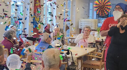 Fasching. Senioren sitzen an Tischen. Durch die Luft wirbelt buntes Konfetti.