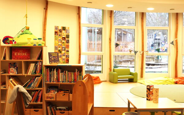 Freizeitraum, mit Regalen mit Büchern. bodengroßen Fenstern, Eine kleine grüne Couch steht vor einem Fenster. Vorne ist ein kleiner Tisch mit Hockern, auf dem Tisch ist ein Jenga Turm / Holz-Stapelspiel