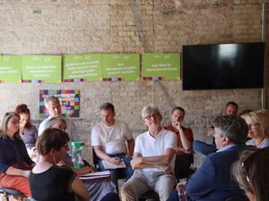 12 Menschen sitzem auf stühlen im Kreis und reden miteinander. Eine Frau hat die grüne Sanduhr auf dem Schoß.