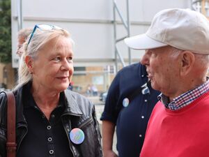 Draußen. Eine Frau und ein Mann sehen sich an. Frau ist blond, die Brille hat sie auf den Kopf geschoben, sie trägt eine schwarze Lederjacke, Der Mann alt, graue Haare lugen unter einem hellen Base Cape hervor, er trägt einen roten Pullover.