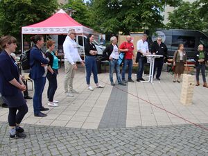 Draußen. 11 Personen stehen in einem Halbkreis um einen Jenga-Turm. Im Hintergrund ein AWO Pavillon und Biertischgarnituren wo ältere Menschen sitzen.