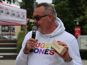 Mann mit Mikro in der rechten Hand und  beschrifteten Holzklotz mit dem Wort: Akzeptanz. Er trägt kurze graue Haare, eine Sonnenbrille und einen weißen Hoody mit bunter Aufschrift.