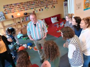 In einem Kita-Raum mit Spielzeug auf dem Fußboden. Die Gruppe Frauen steht im Halbkreis um einen Mann. Mann ist etwas älter, kurze Haare, Brille. Er trägt ein bunt längsgestreiftes helles Hemd.