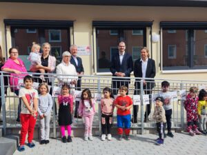 11 Kinder stehen vor der Rampe vor dem Gebäude. 7 Erwachse, darunter die drei Männer im Anzug stehen verteilt und locker auf der Rampe am Gebäude.