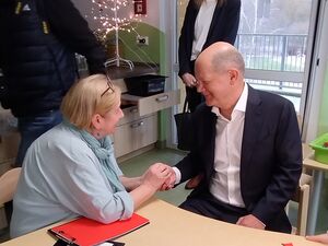 Eine Frau und ein Mann sitzen an einem Tisch. Die Frau hat beide Hände um die Hand des Mann gelegt. Mann trägt die Anzugjacke offen und ein weißes Hemd darunter. Im Hintergrund steht eine Frau vor dem Fenster.