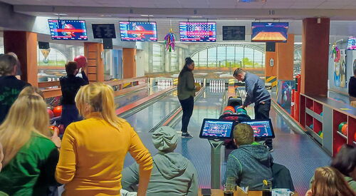 Eine Bowlinghalle. Es wird gebowlt, einige sitzen am Tisch mit Getränken und schauen zu.