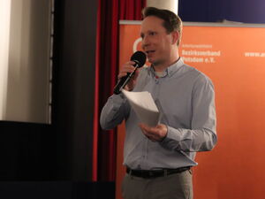 Moderator steht mit Notizen und Mikro vor der Kinoleinwand. Er trägt ein helles Hemd und graue Hose.