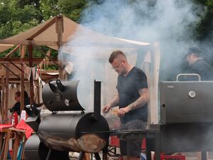 Mann mit Undercut, Brille, schwarzes T-Shirt und Tatowierungen an den Armen steht an einem großen Grill. Es qualmt sehr.