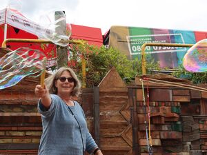 Eine Frau macht große Riesen-Seifenblasen mit einem Seifenblasen-Set. Die Frau ist blond, trägt eine Sonnenbrille und ein blaugraues T-Shirt.