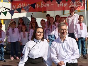 Kinderchor auf der Bühne. Mädchen und Jungen lächeln in die Kamera. Vor der Bühne stehen eine Frau und ein Mann. Beide tragen lange weiße Hemden mit schwarzen Knöpfen.