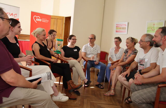 10 Menschen sitzen in einem Raum auf Stühlen. Eine Frau hat eine grüne sanduhr auf dem Schoß, die andere sitzen sich gegenüber.
