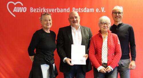 Portrait: 4 Personen vor roter AWO Wand. Frau, blonde lange Haare hochgesteckt, Brille auf den Kopf geschoben, Mann mit Glatze, weißem Hemd, schwarzem Sakko hält ein Papier vor sich, Frau mit weißen Haaren und Brille und großer schlanker Mann mit Brille.