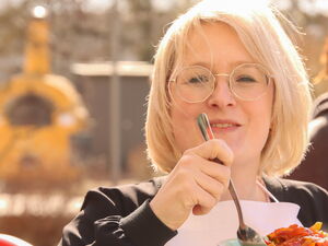 Blonde Frau mit Brille und weißer Schürze hält einen gefüllten Teller und Löffel. Sie lächelt in die Kamera.