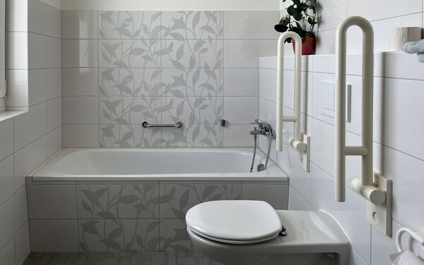 Badezimmer: Wanne mit Haltegriff an der Wand. WC mit hochgeklappten Haltegriffen rechts und links neben dem WC montiert.