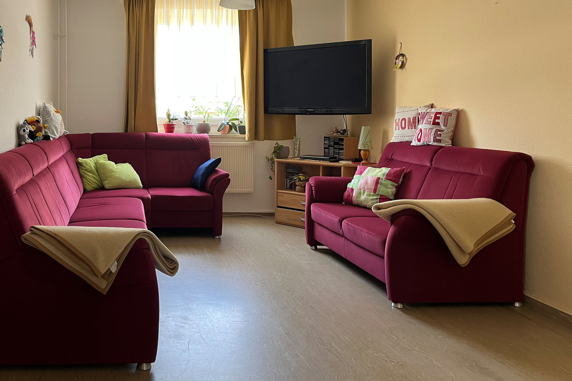 Gemütlicher Gemeinschaftsraum, mit einer großen Echcouch und gehgenüber eine Couch. Beide Möbel sind Weinrot. In der rechten hinteren Ecke ist ein TV an der Wand angebracht.