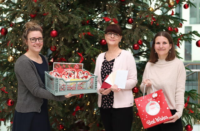 Drei Frauen vor einem großem geschmücktem Weihnachtsbaum mit roten Kugeln und Schleifen. Zwei Frauen tagen eine Faltbox mit Herzen drin. Die dritte hat eine rote Weihnachtstüte mit Aufschrift: Best Wishest